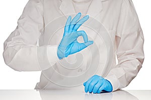 Female doctor in white medical gown and blue sterilized surgical gloves showing OK sign