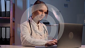 Female doctor working with laptop computer and typing information from paperwork. Hospital night background.