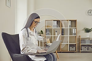 Female doctor working with laptop while sitting in chair on office interior background.