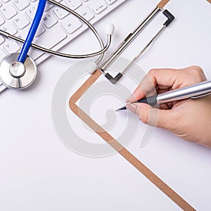 Female doctor writing a medical record case over clipboard on white working table with stethoscope, computer keyboard. Top view,