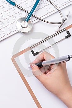 Female doctor writing a medical record case over clipboard on white working table with stethoscope, computer keyboard. Top view,