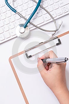 Female doctor writing a medical record case over clipboard on white working table with stethoscope, computer keyboard. Top view,