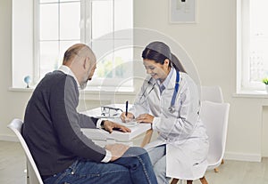 Female doctor writing medication order for senior patient in clinic. Elderly man on visit to his healthcare provider