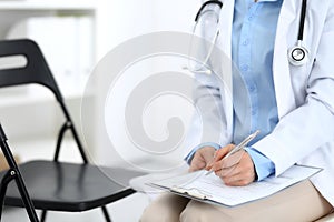 Female doctor writing up medication history records form on clipboard, while sitting at the chair. Physician at work in