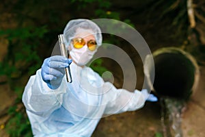 Female ecologist or epidemiologist demonstrates a test tube with water, second hand pointing to a sewer coming out of the ground