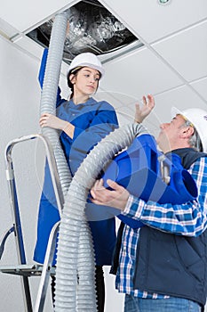 Female electrician installing ac pipe in ceiling with master worker