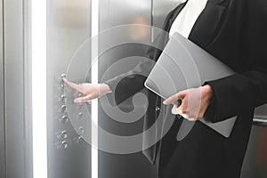 Female employee pressing the elevator button and holding the laptop. Businesswoman in the formal black suit is pushing the button