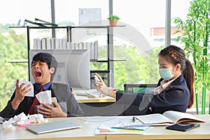 Female employee wearing medical face mask and spraying alcohol spray to her male colleague who is coughing and sneezing