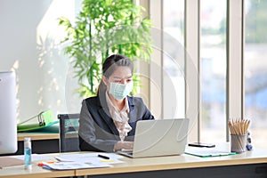 Female employee wearing medical face mask while working alone because of social distancing policy in the business office