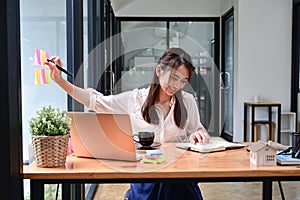 Female employee working with laptop computer and making note on sticky notes on glass wall in office.