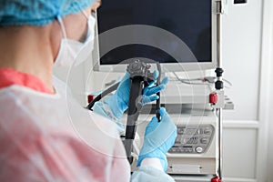 A female endoscopist surgeon in a protective suit, cap, mask and gloves holds an endoscope in front of a monitor