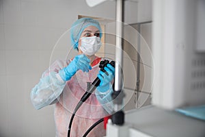 A female endoscopist surgeon in a protective suit, cap, mask and gloves holds an endoscope in front of a monitor