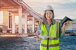 Female engineer with computer and blueprint is standing infront City tranportation bridge over river for city development