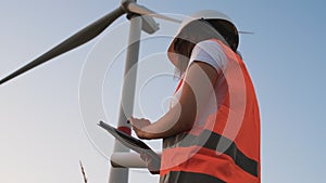 Female an engineer in a helmet and an orange vest controls the operation of a wind turbine using a tablet on the sky