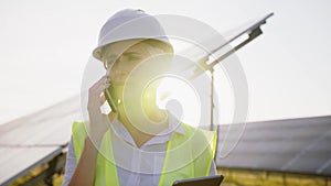 Female engineer holding tablet in her hand and talking on phone. Business woman working remotely among rows of solar