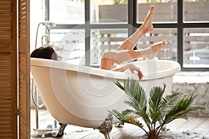 A female enjoy lies in a white bathtub and looks out the street through a large window. Back view shot