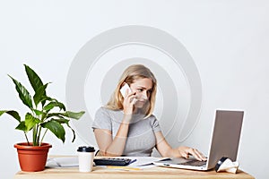 Female enterpreneur, being busy with work, keyboarding on laptop computer, calling her business partner while consulting something