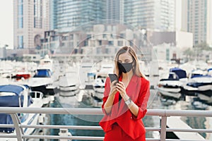 Female entrepreneur standing at city business center in front of luxury yachts and skyscrapers and looking at smartphone screen