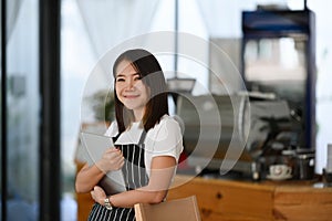 A female entrepreneur wearing black apron hands holding tablet and standing in her own coffee shop.