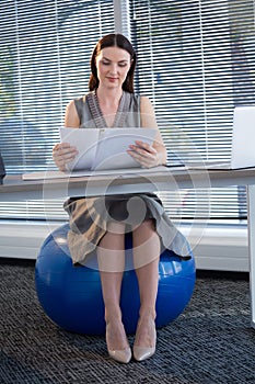 Female executive sitting on exercise ball while reading documents at desk