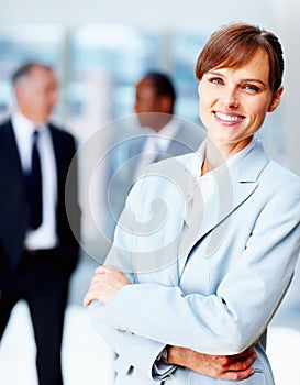 Female executive standing casually with colleagues in background. Business woman smiling while standing with arms folded