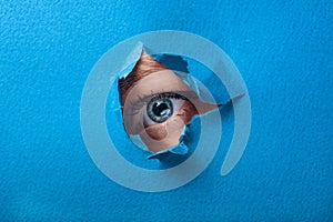 A female eye looks through a hole in a paper blue background