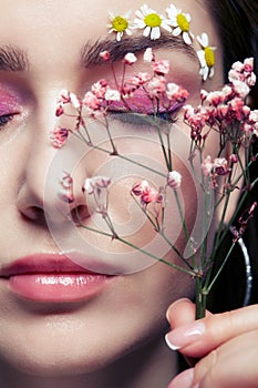 Female face with pink makeup. Woman with Gypsophila flowers in hand near face. Brows are decorated by small chamomile flowers