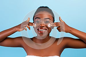 Female facial skincare. African american lady applying moisturizer cream on cheeks, posing over blue studio background