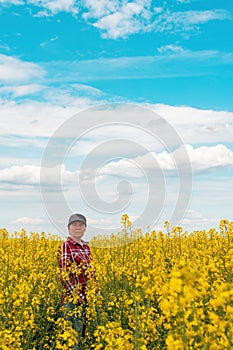 Female farm worker wearing red plaid shirt and trucker`s hat standing in cultivated rapeseed field in bloom and looking over crop