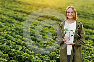 Female farmer holds in hands soybean plant with health leaves and roots, looks at camera at soya field. Agronomist