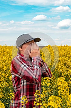 Female farmer with pollen allergy sneezing in blooming rapeseed field. Farm worker wearing plaid shirt and trucker`s hat with