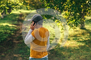 Female farmer using smartphone in walnut tree orchard. Farm worker wearing orange t-shirt and trucker`s hat with mobile phone in