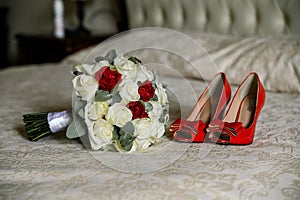 Female fashion red wedding shoes with bride`s bouquet of white and red roses lie on the bed. Close up