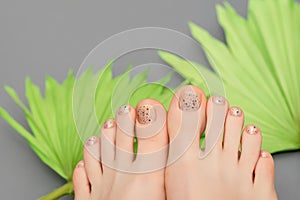 Female feet with beige nail design. Glitter beige nail polish pedicure with brown nail art on green tropic leaves background