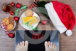 Female feet on digital scales with sign omg! surrounded by Christmas decorations, bottle, glass of alcohol and sweets.