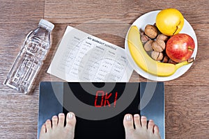 Female feet on digital scales with word ok surrounded by bottle of water, plate with healthy food and workout schedule paper.