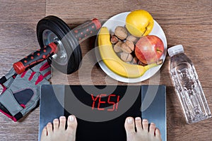 Female feet on digital scales with word yes surrounded by sport accessories AB roller wheel, fitness gloves, healthy food.