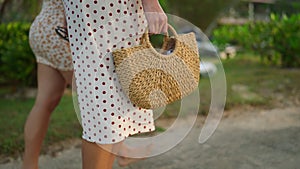Female feet in dresses going barefoot to tropical beach along path. Two young women walking, holding straw bag on exotic