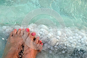 Female feet with manicure on stone, covered with salt formations between the waves in the water. Crystallization of salt on stone