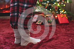 Female feet in pajama and winter socks standing near a Christmas tree with gifts and fireplace at morning. Concept.