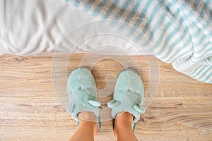 Female feet in soft blue slippers on a wooden floor near the bed close-up. Concepts of cozy fleece home shoes and warm domestic