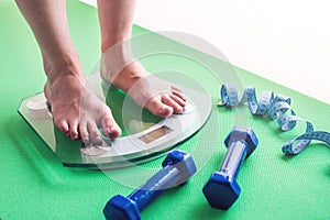 Female feet standing on electronic scales, dumbbells and measuring tape. Concept of slimming and weight loss