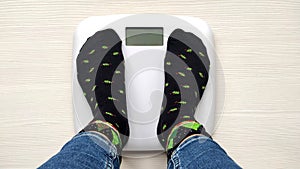 Female feet standing on electronic scales for weight control in funny New Year socks on white wooden background