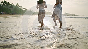 Female feet walking barefoot in sea water edge at sunrise. Foamy waves, reflections on water. Two attractive girls going