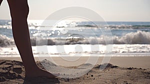 Female feet walking at the sea beach on a sunny day with waves at background. Legs of young woman stepping at the sand