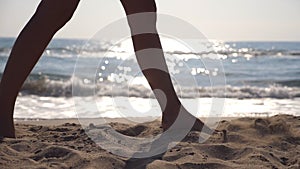 Female feet walking at the sea beach on a sunny day with waves at background. Legs of young woman stepping at the sand