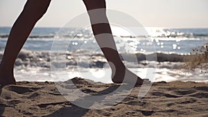 Female feet walking at the sea beach on a sunny day with waves at background. Legs of young woman stepping at the sand