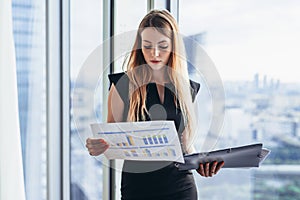 Female financial analyst holding papers studying documents standing against window with city view