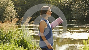 Female with fitness mat walking in summer park. Woman strolling in park along riverside, going at outdoors yoga class. Meditation