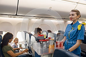 Female flight attendant serving food to passengers on aircraft. Hostess walking with trolley on aisle